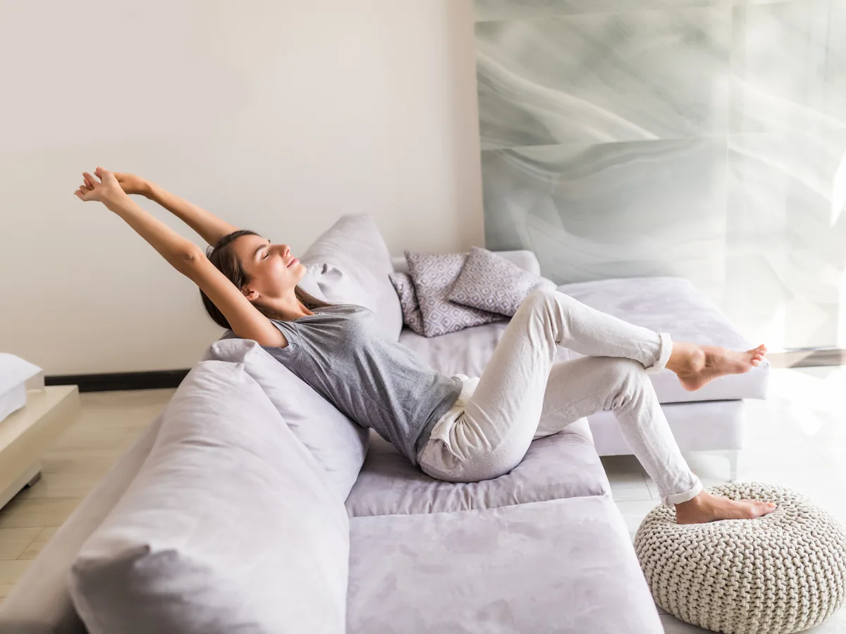 Young woman stretching with her arms overhead and relaxing on a sofa in a bright, airy living room